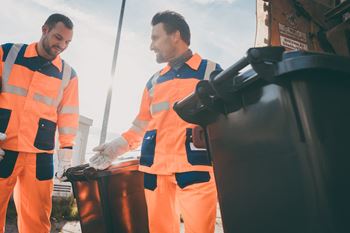 Two garbage men dressed in bright colored uniforms working with two garbage cans